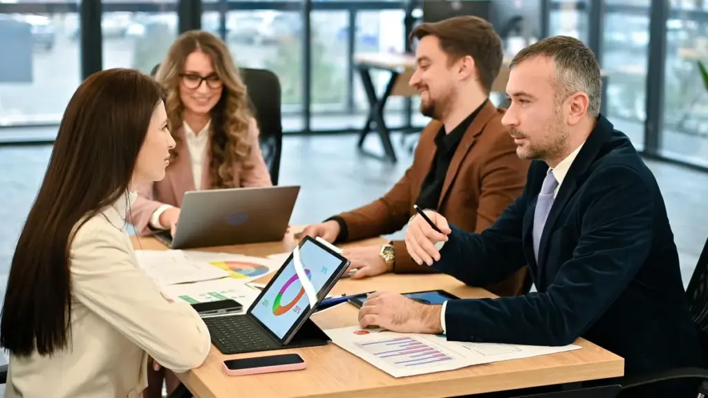 Quatro profissionais conversam em uma mesa de reunião com notebook, tablet e gráficos impressos