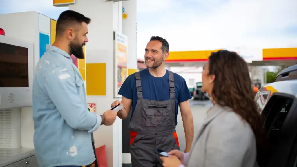 Frentista conversa com clientes em um posto de combustível; um deles segura um cartão